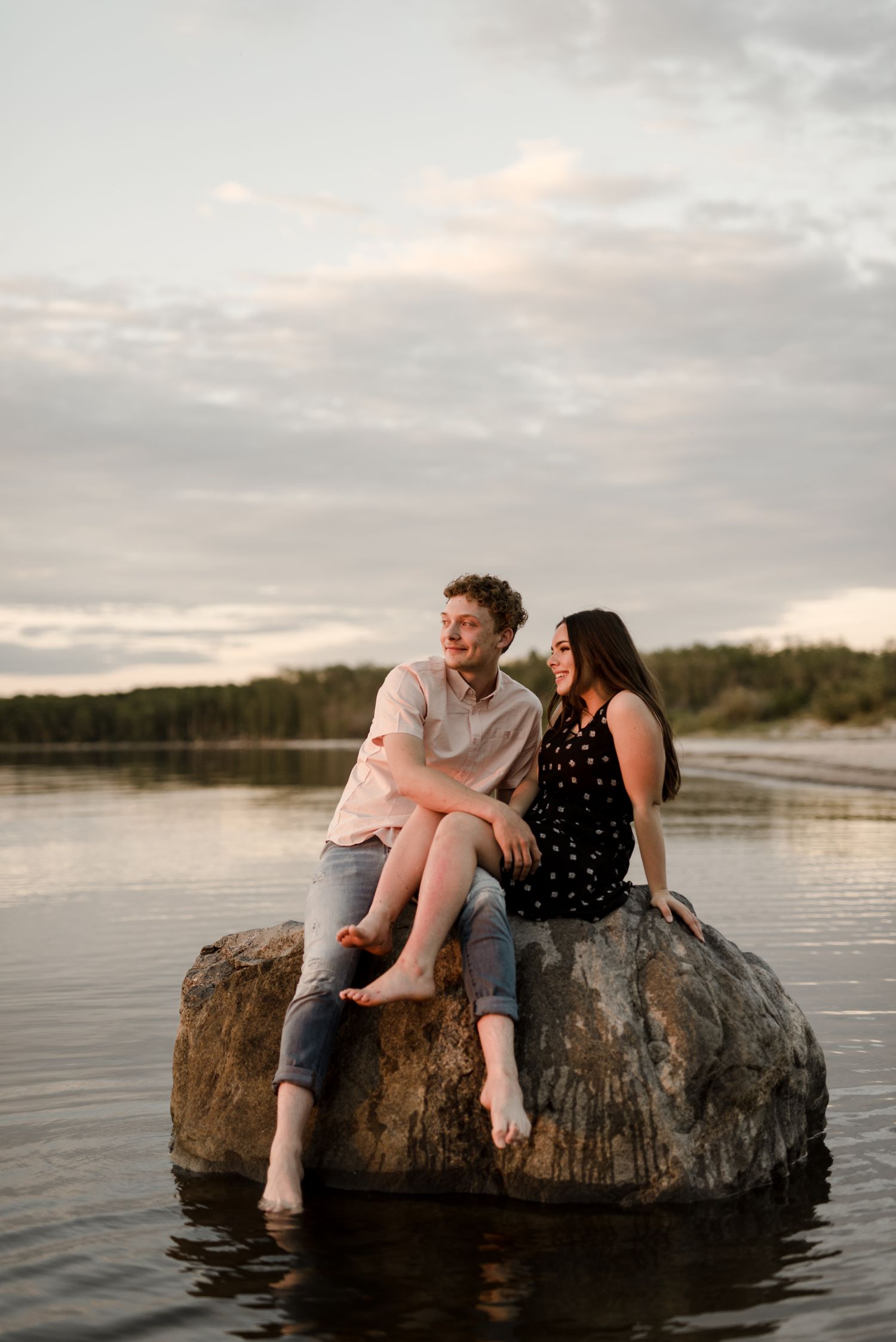 Couple sitting on a rock in the water at their Grand Beach engagement session in Manitoba. Beautiful deep pink sunset, photos taken by Vanessa Renae Photography